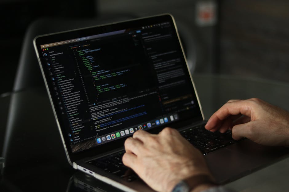 A pair of hands typing on a MacBook laptop with a dark screen displaying lines of colourful code, positioned on a black surface in an indoor setting. The laptop is open, showing a taskbar with applica
