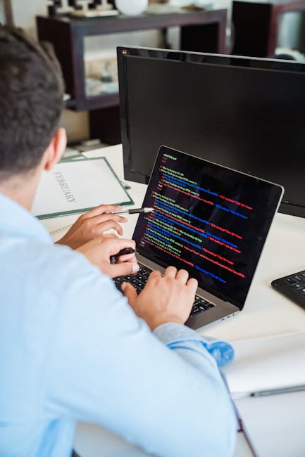 A pair of hands typing on a MacBook laptop with a dark screen displaying lines of colourful code, positioned on a black surface in an indoor setting. The laptop is open, showing a taskbar with applica